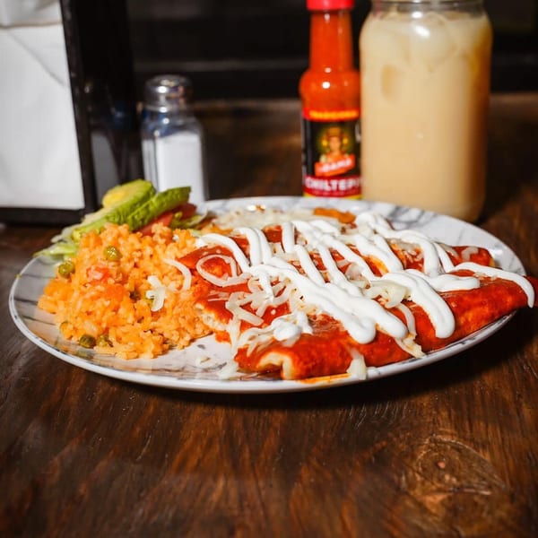 A plate of enchiladas with rice and salad, hot sauce, and a drink on a wooden table.