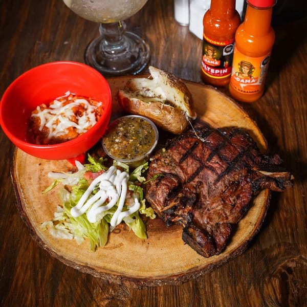 Grilled steak with baked potato, beans, salad, salsa, and hot sauce on a wooden board.