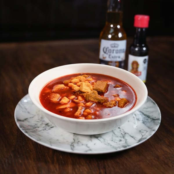 A bowl of red pozole with meat and hominy, a Corona beer, and hot sauce.