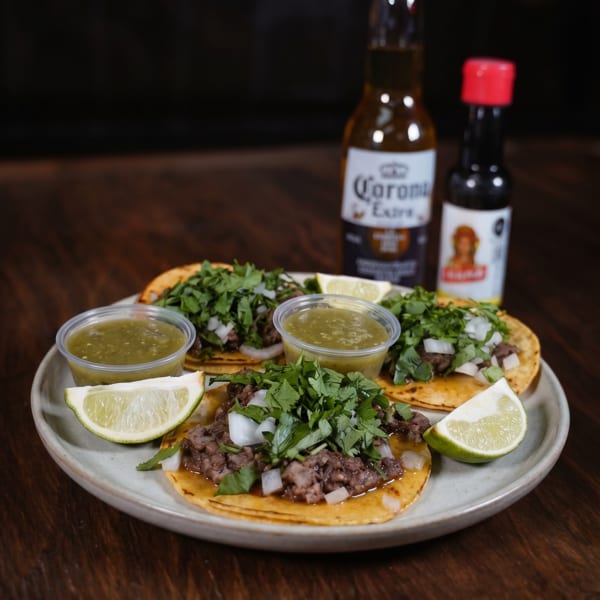 Three street tacos with meat, onions, and cilantro, served with salsa, lime, and a Corona beer.