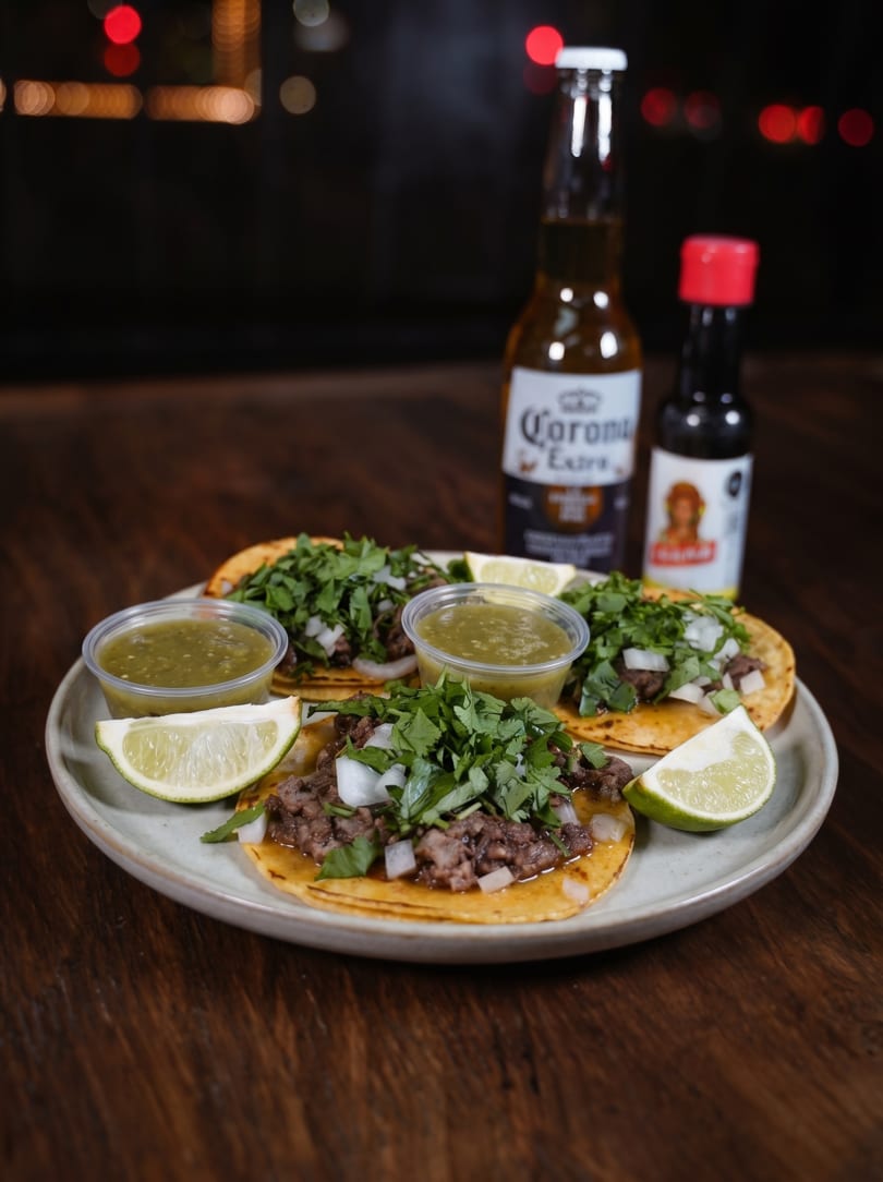 Three street tacos with meat, onions, and cilantro, served with salsa, lime, and a Corona beer.