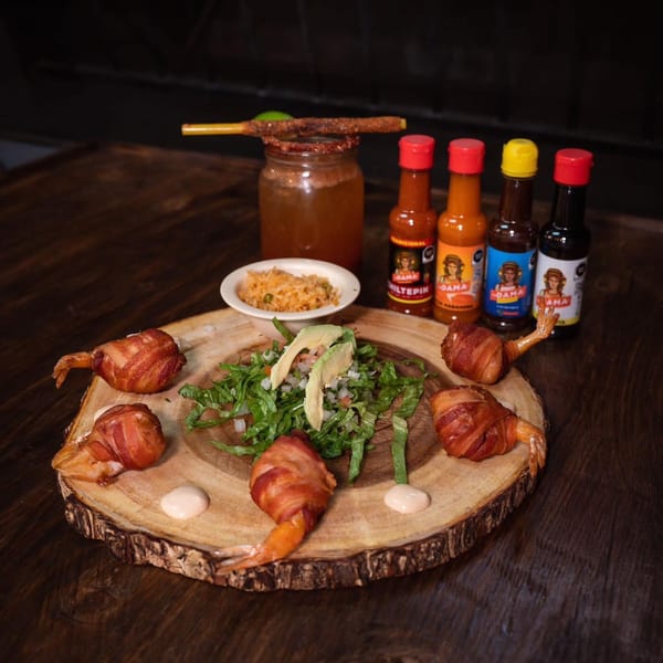 Bacon-wrapped shrimp, salad, rice, hot sauces, and a michelada on a wooden board.